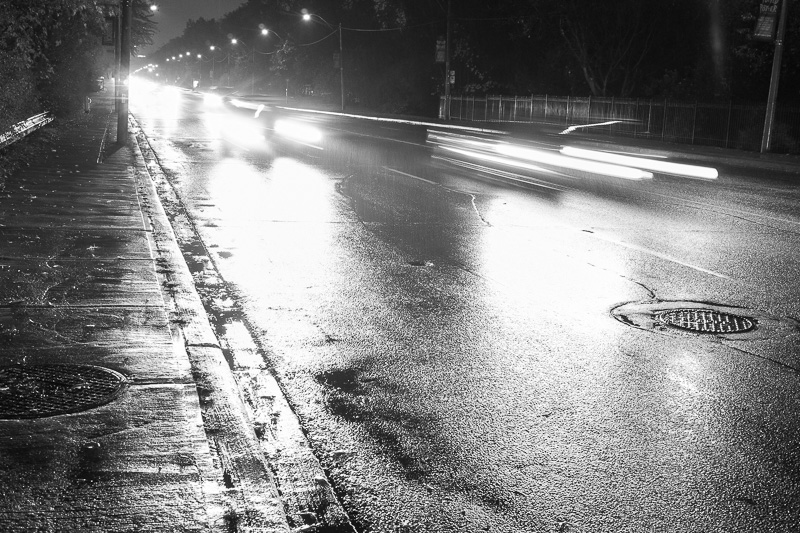 Black and white photograph of car at night passing on wet pavement