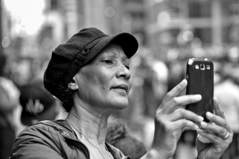 Street photography that captures the details of a woman's face as she takes a photo with her smart phone.