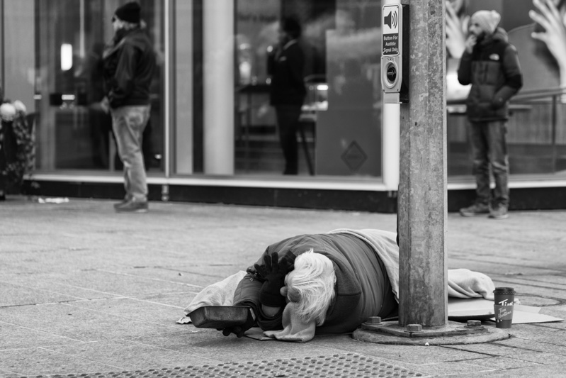 Homeless man lying on the street holding out a tin pan.