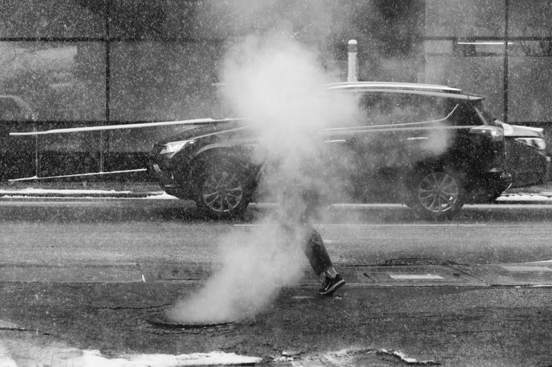 Woman is all but completely obscured by steam from a vent while walking along a sidewalk.