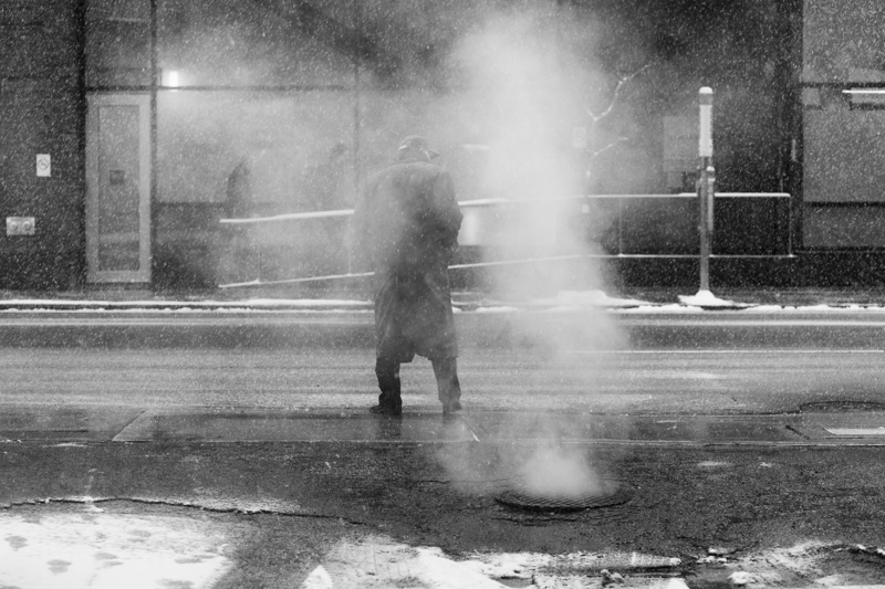 Man waits to cross street while enshrouded in steam.