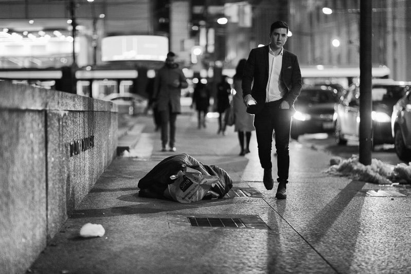Walking south on Bay Street in December, a young man in suit jacket passes a homeless man lying on a grate.
