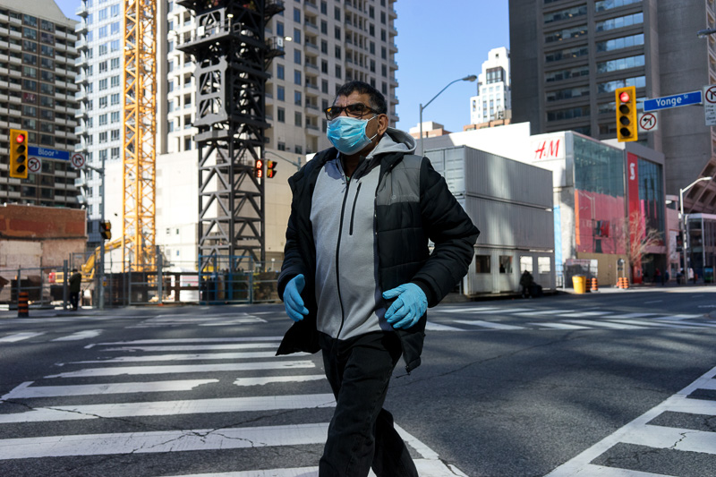 A man wearing a blue face mask and blue gloves crosses the Bloor/Yonge intersection in Toronto.