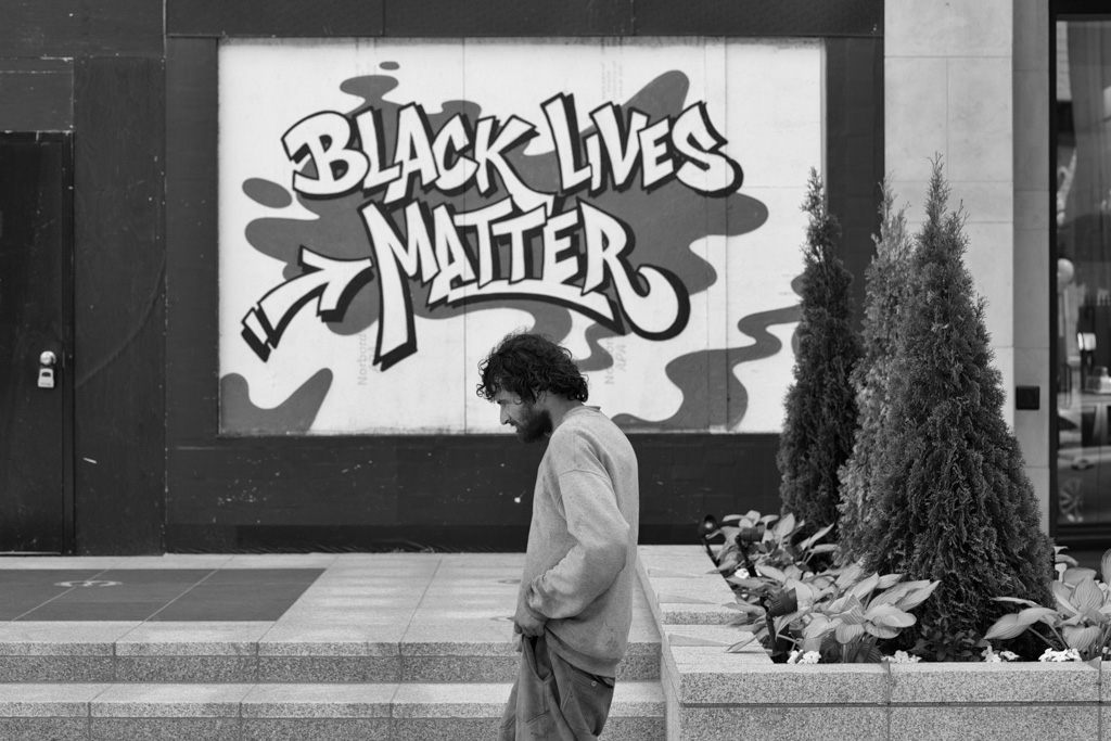 Man passes shop window covered by plywood spray painted with the message: Black Lives Matter.