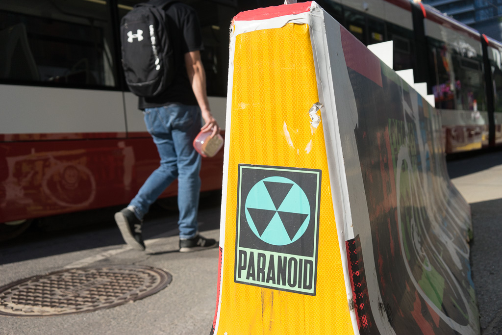 A poster that says "paranoid" stuck to the reflective yellow end of a concrete barrier at a streetcar stop.