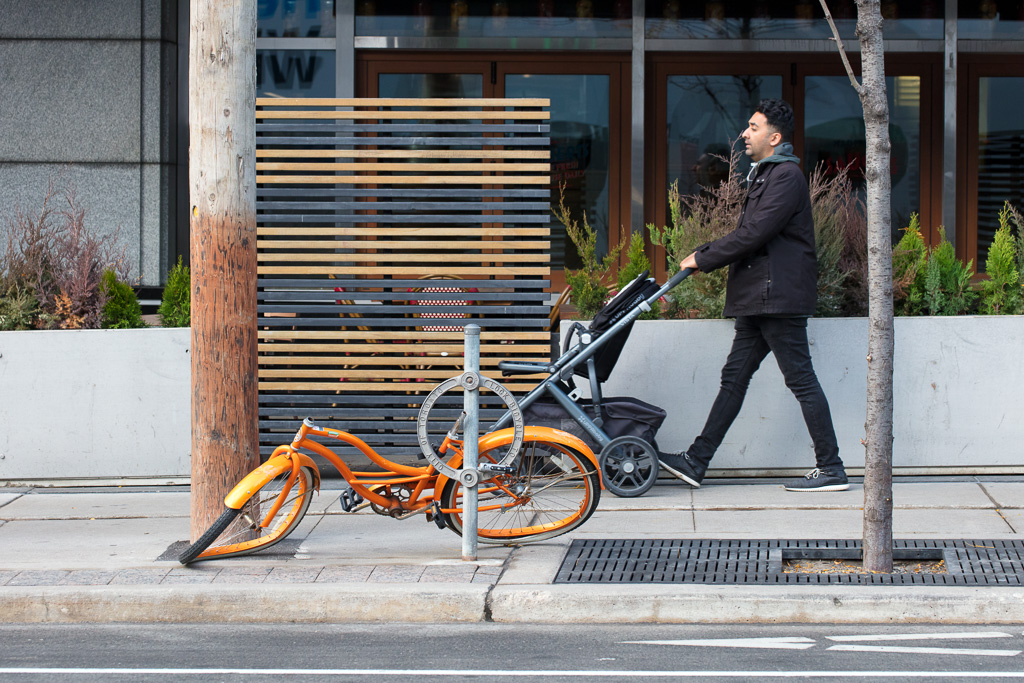 Man pushes empty baby stroller past broken down bicycle.