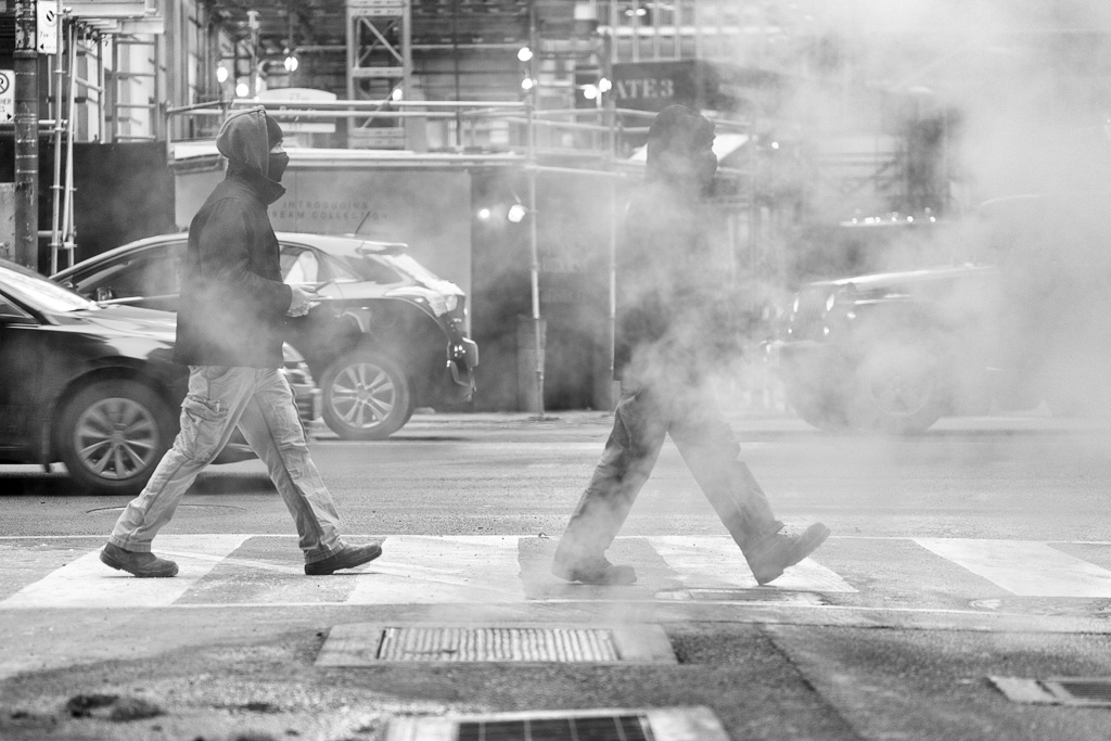 Two men cross the street behind a steam vent.