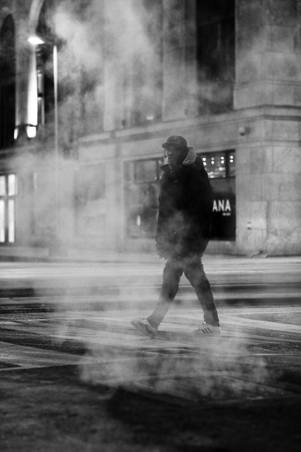 Man cross the street at night behind a steam vent.