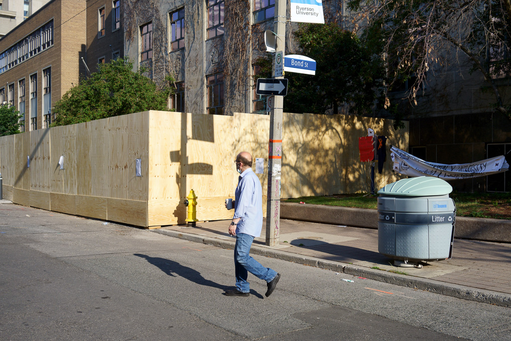 Man walks past plywood enclosing the site of the former statue of Egerton Ryerson.