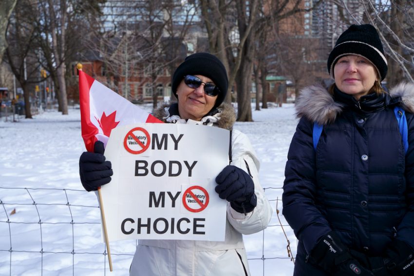 Winter scene in Queen's Park, Toronto. Two women pose on a snow-covered lawn. One of the women holds a Canadian flag and a sign that says "My Body My Choice" and the crossed out words "Mandatory Vaccines"